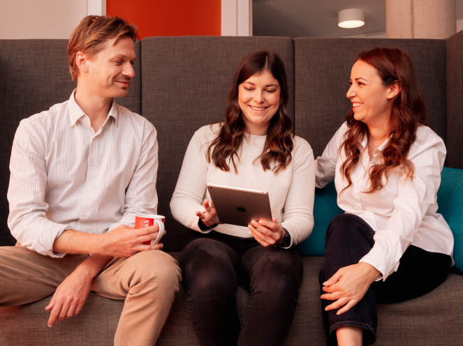Drei Personen sitzen auf einem Sofa, lächeln und schauen auf ein Tablet.