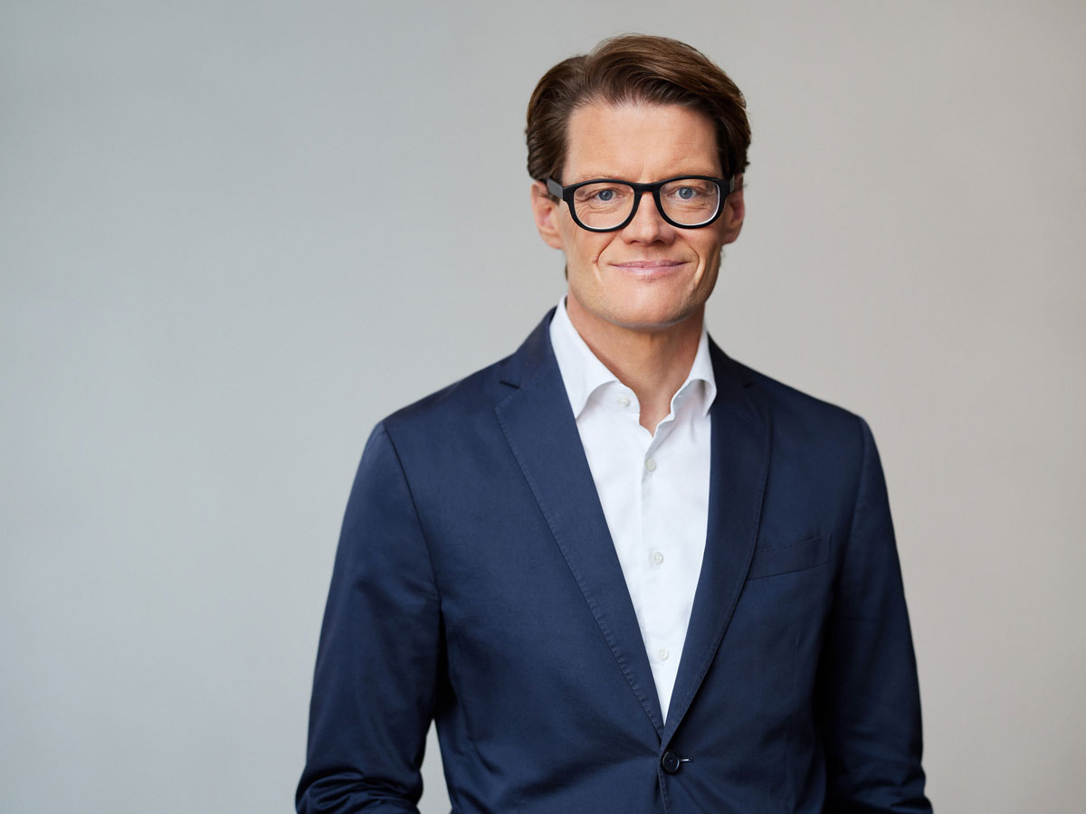 A person in a navy suit and glasses smiles against a plain background.