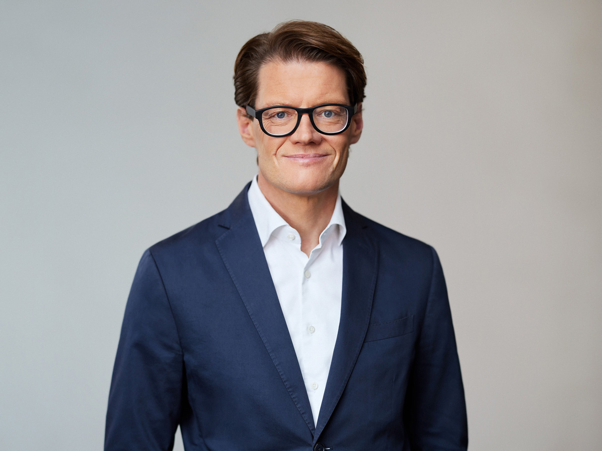 Ein Mann in Anzug und Brille lächelt vor neutralem Hintergrund.