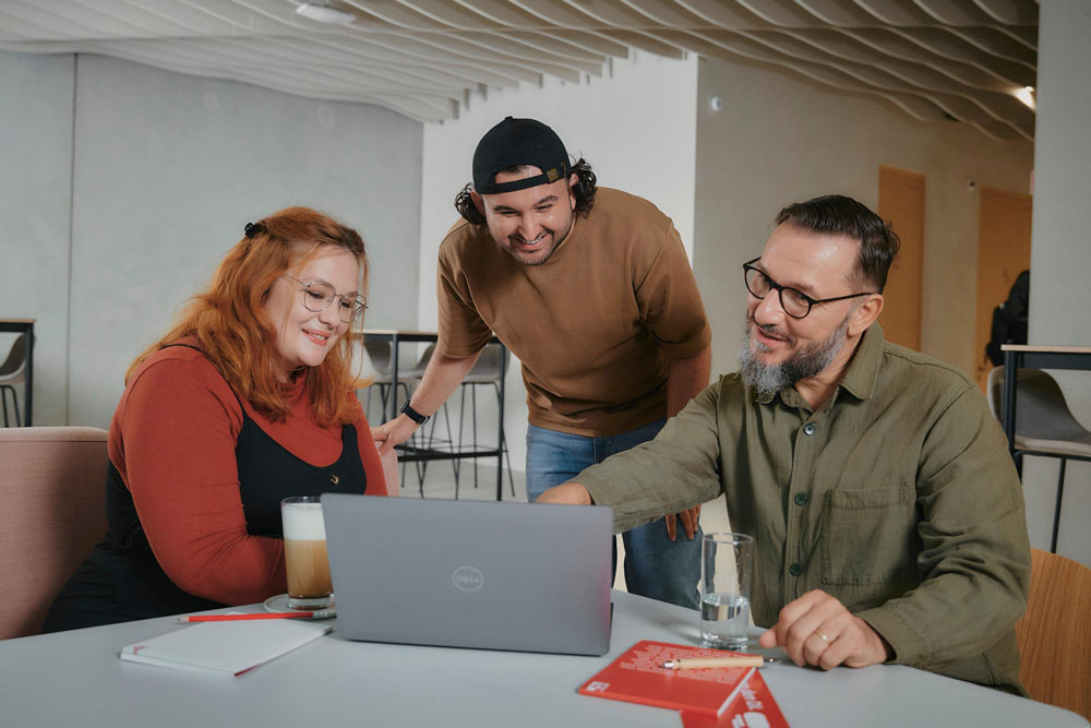 Drei Personen lächeln und schauen auf einen Laptop in einem modernen Büro.