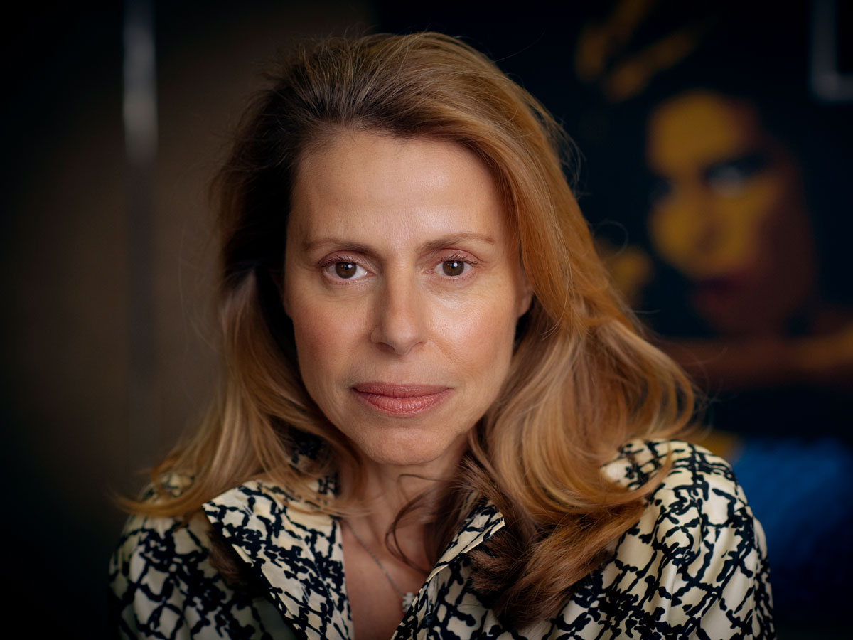 A woman with long hair in a patterned blouse sits in front of a blurred dark background.