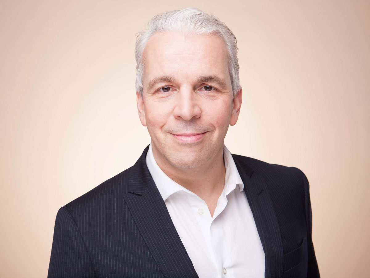 A person with gray hair in a dark suit and white shirt smiles against a beige background.