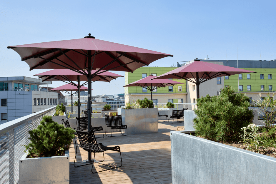 Dachterrasse mit Holzboden, roten Sonnenschirmen, schwarzen Stühlen und grünen Pflanzen in Metallkästen.