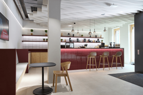 Moderne Café-Bar mit roter Theke, drei Holzstühlen, hängenden Lampen und Pflanzenregalen an der Wand.