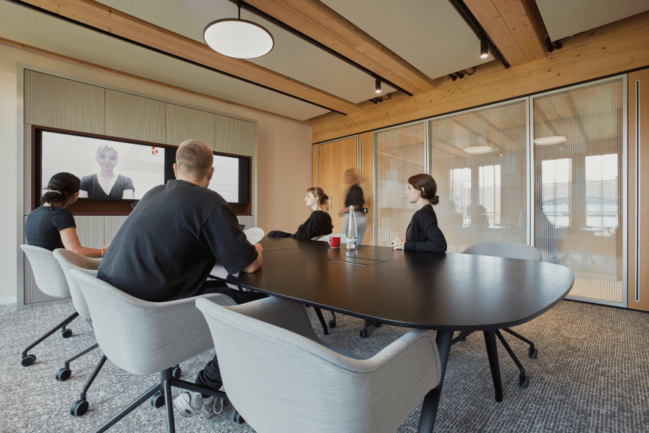 Fünf Personen in einem Konferenzraum, schauen auf einen großen Bildschirm mit Videokonferenz.
