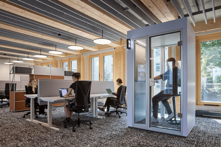 Offenes Büro mit Holzelementen, mehreren Personen an Schreibtischen und einer Person in einer Telefonkabine.