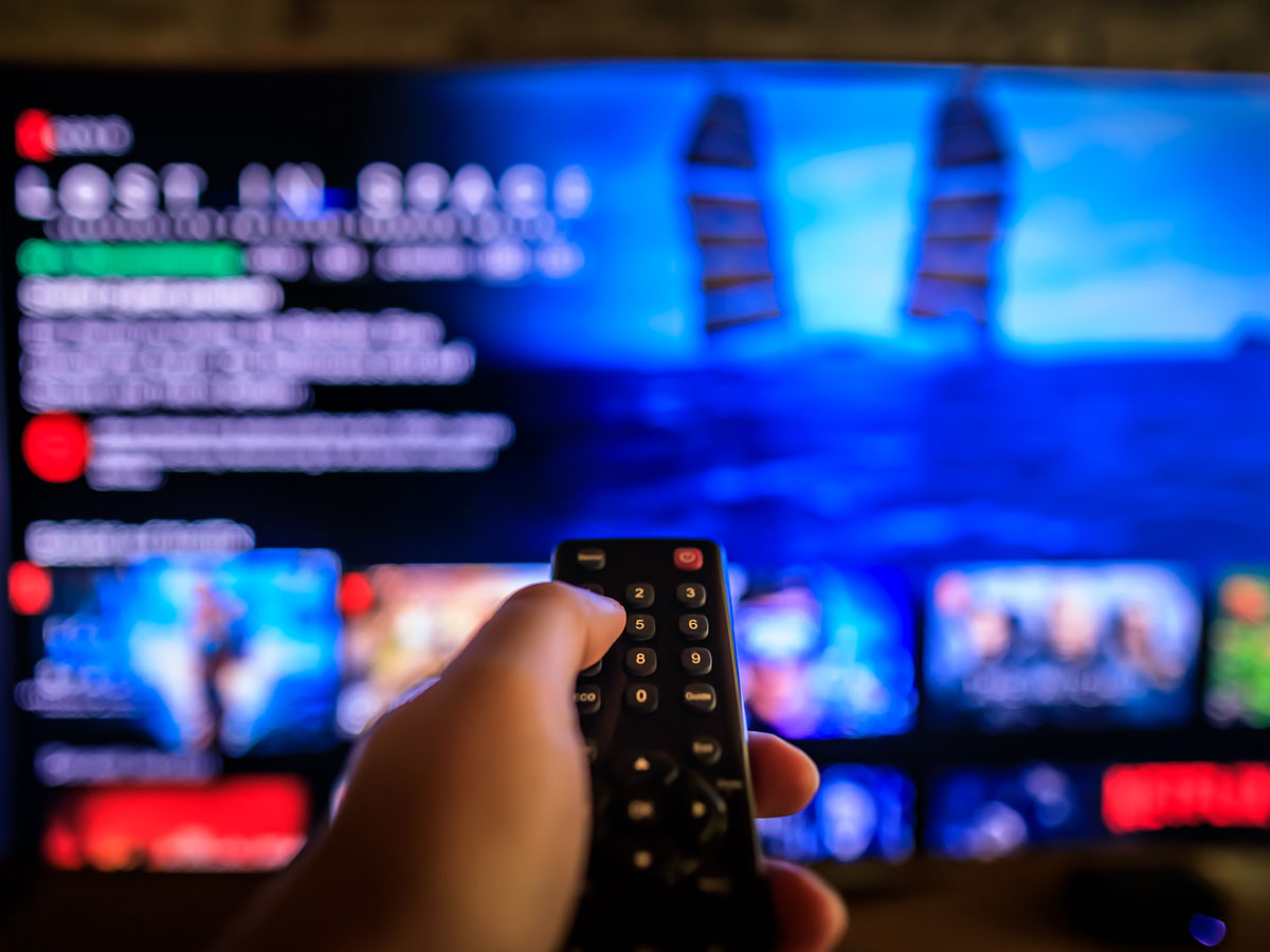 A hand holds a remote control in front of a TV displaying a blurred streaming service interface.