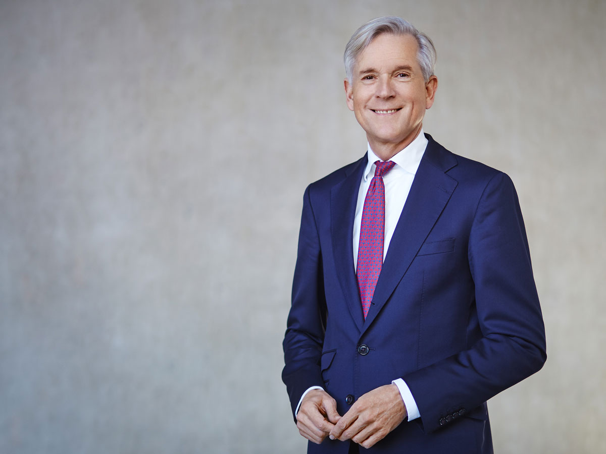 A man in a blue suit and red tie smiles against a plain gray background.