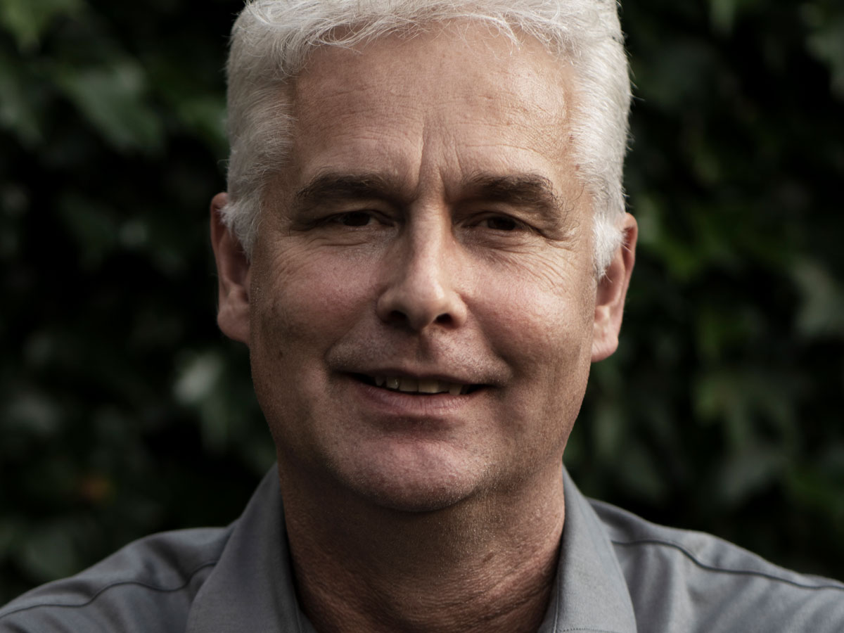 A man with short gray hair smiles slightly, wearing a gray shirt, with a blurred green leafy background.