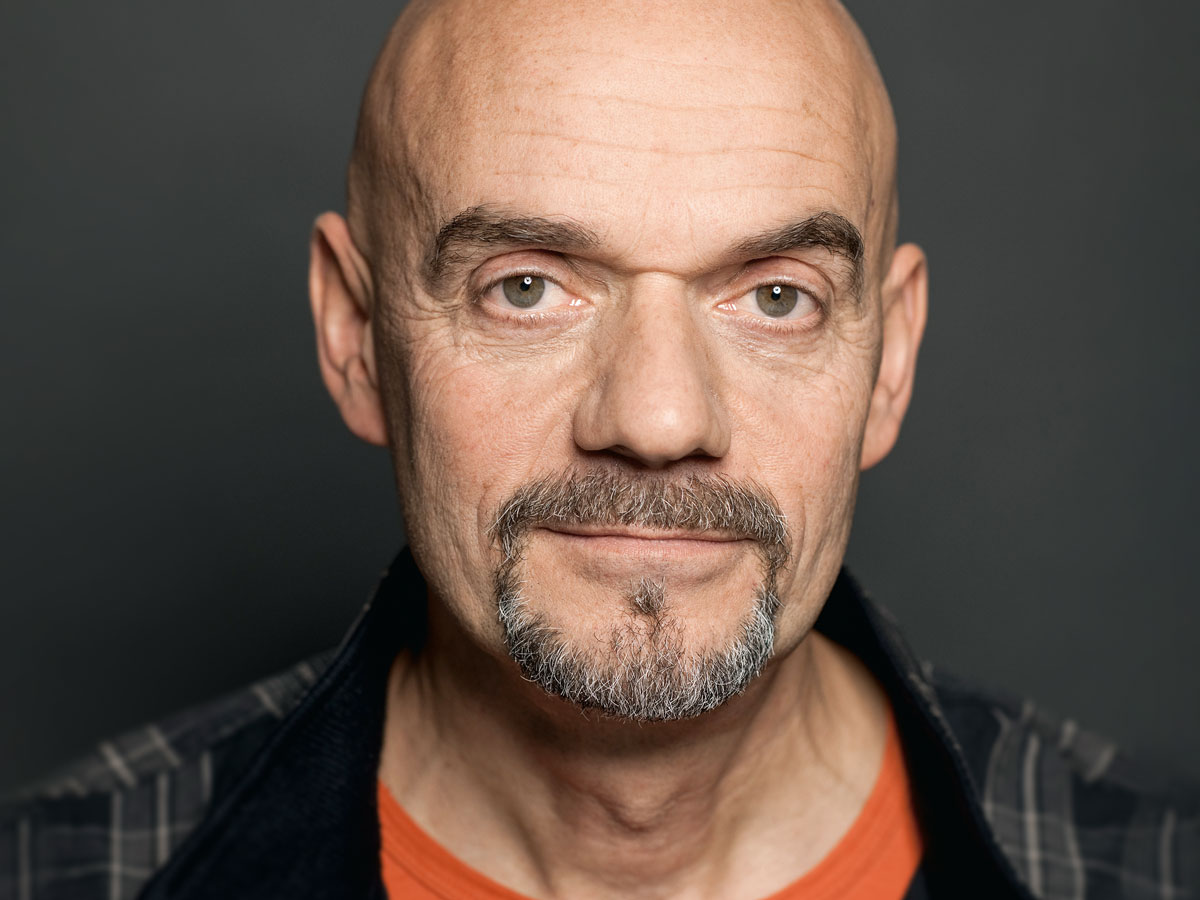 A bald man with a goatee and plaid shirt looks directly at the camera against a gray background.