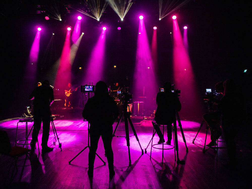 Silhouetted camera crew films a band on stage under vibrant purple spotlights.
