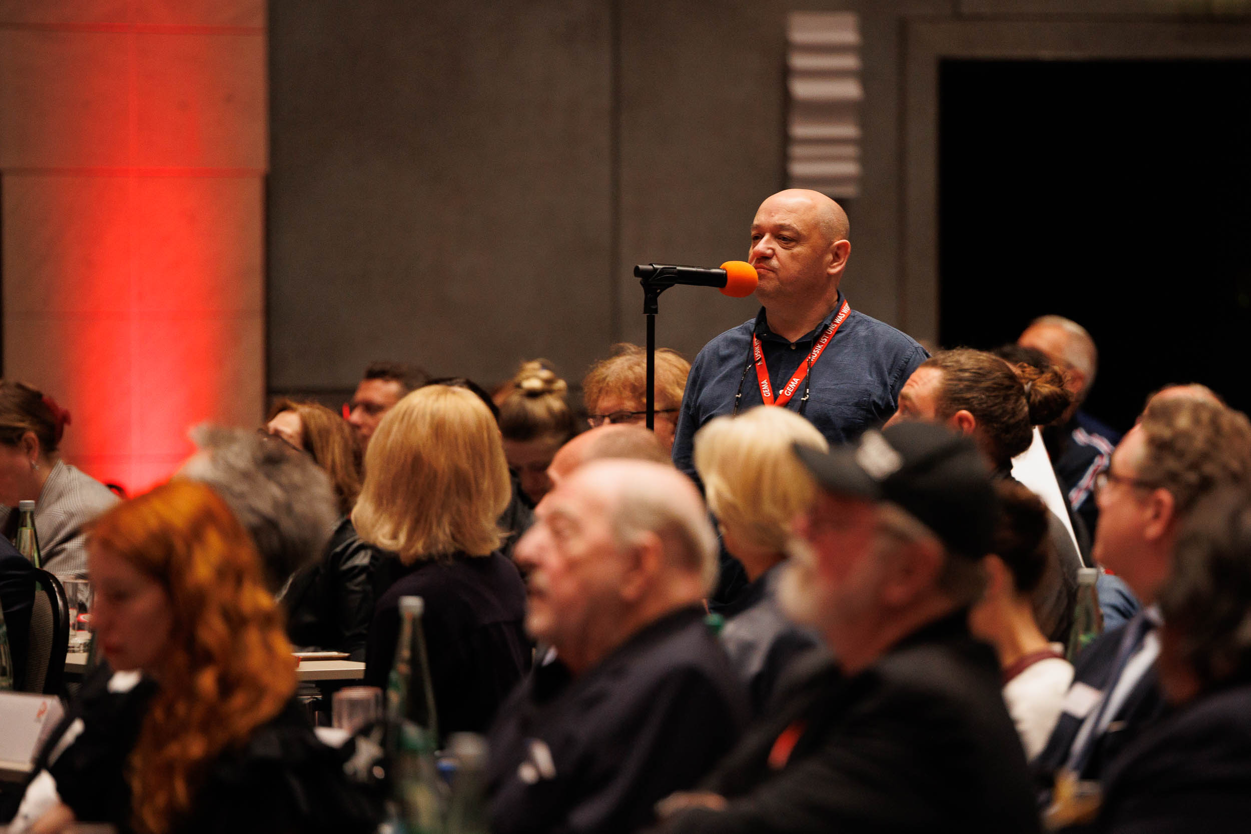 A man speaks into a microphone with an orange cover at a crowded conference, surrounded by seated attendees.