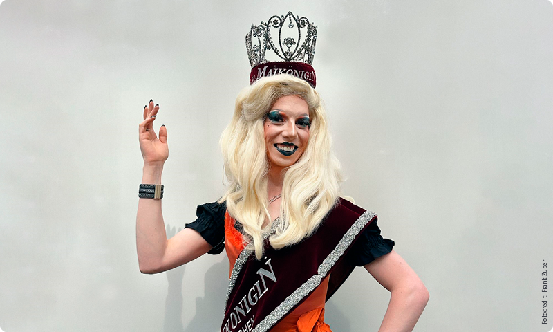Person in a sash and crown with long blonde hair, waving and smiling against a plain background.