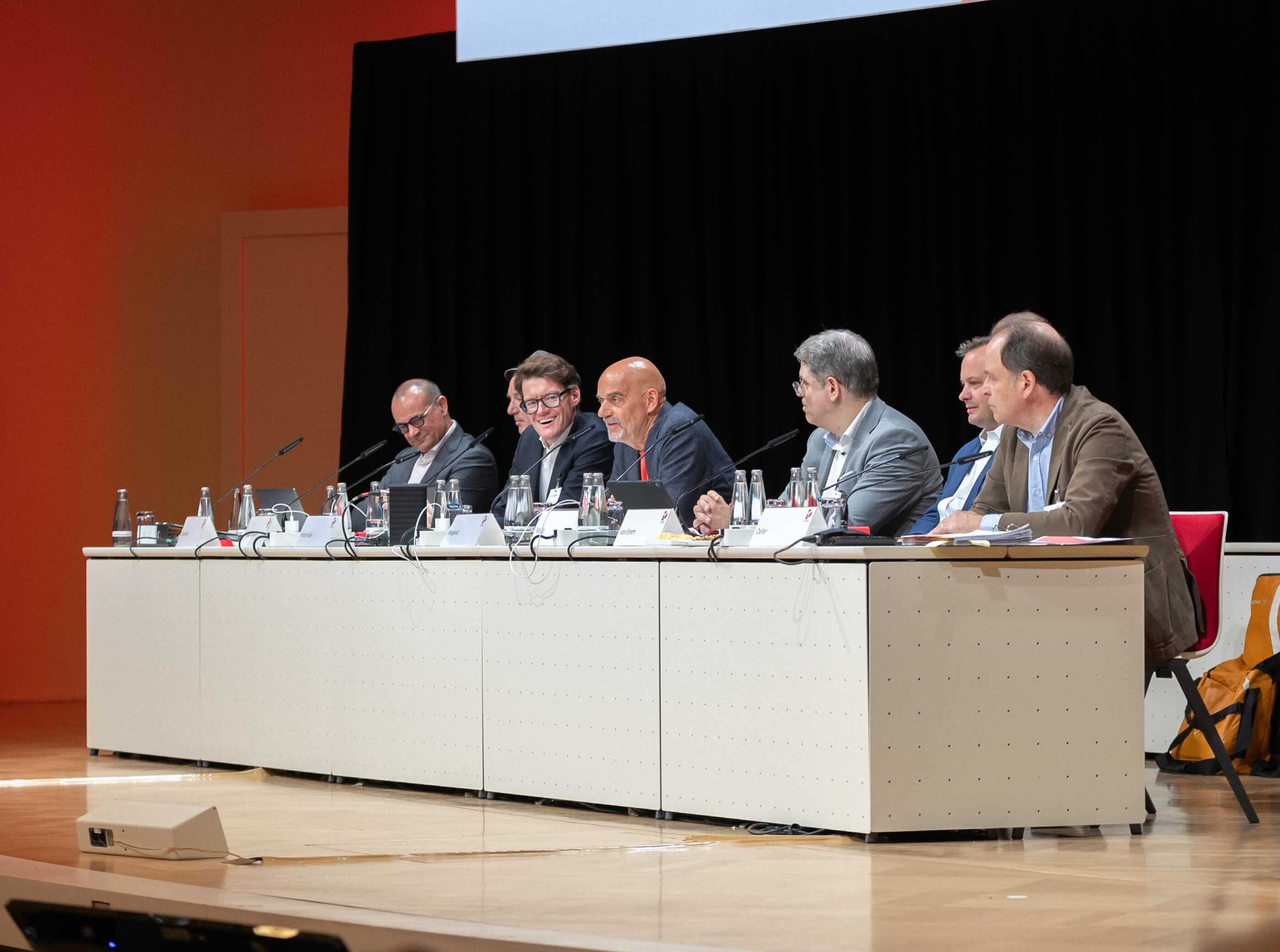 Sieben Männer sitzen bei der Mitgliederversammlung an einem Konferenztisch auf der Bühne.