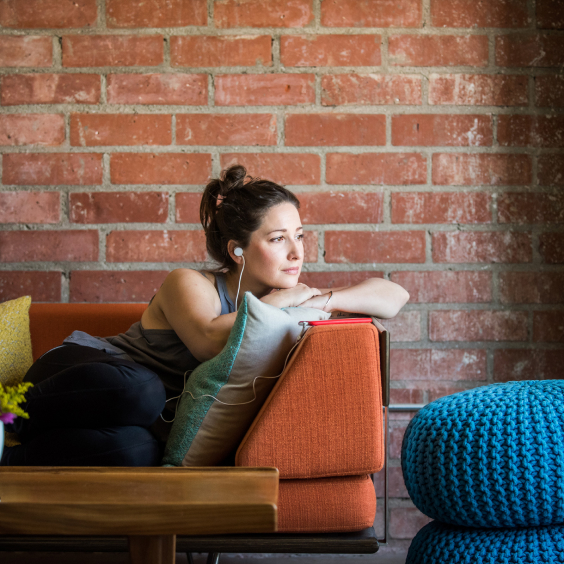Woman with earbuds lounges on an orange sofa, leaning on pillows, against a brick wall, with a blue pouf nearby.