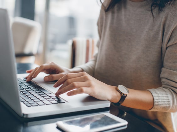 Person typing on a laptop at a table, smartphone nearby, wearing a watch and a beige sweater.