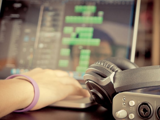 A hand on a laptop keyboard, wearing a purple wristband, with headphones and audio equipment nearby.