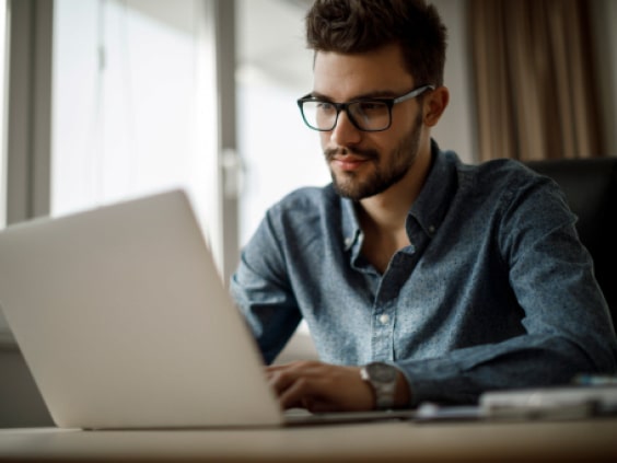 A man in glasses and a blue shirt works on a laptop at a desk near a window.
