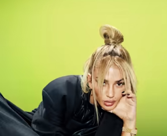 Person with blonde hair in a bun, wearing a black outfit, poses against a bright green background.