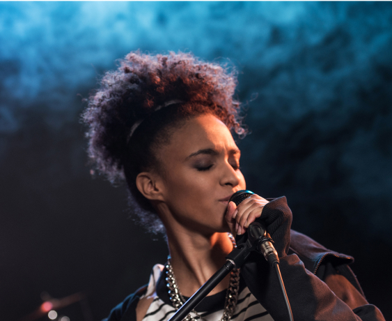 A person with curly hair sings passionately into a microphone on a dimly lit stage with blue smoke in the background.