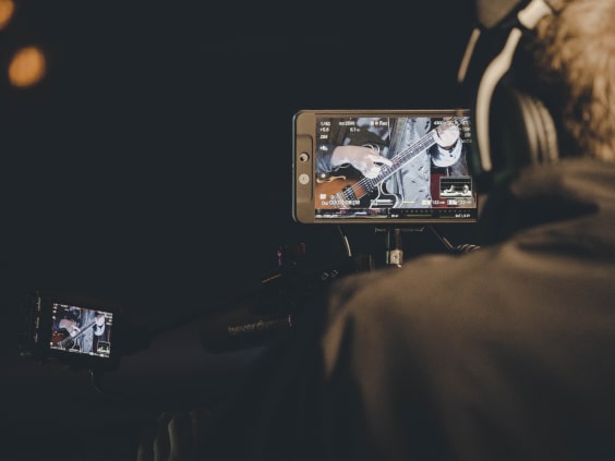 Person filming a guitarist on a camera, screen shows close-up of guitar and hands in a dimly lit setting.