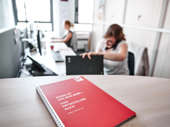 Gema Buch liegt auf einem Tisch. Im Hintergrund sind zwei Frauen zu sehen, die am Schreibtisch sitzen und arbeiten.