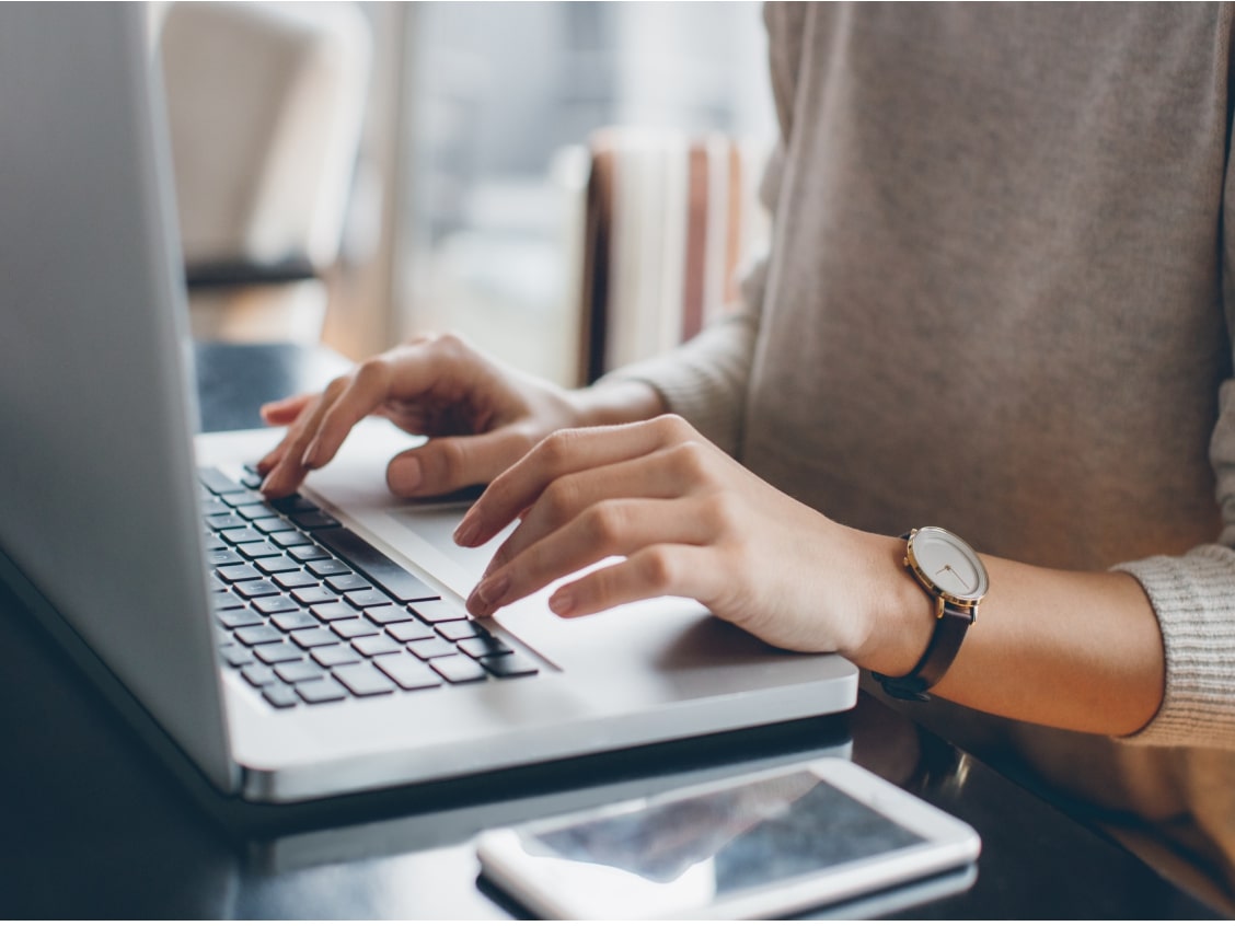Hands typing on a laptop keyboard, with a smartphone and a watch visible on the desk.