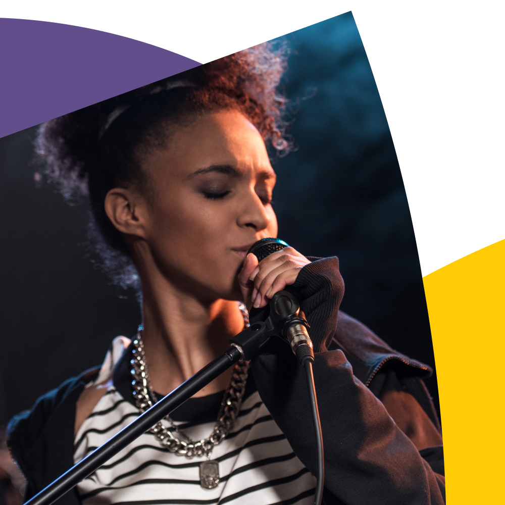 Singer in striped shirt and jacket passionately sings into a microphone on stage, with colorful abstract shapes in the background.