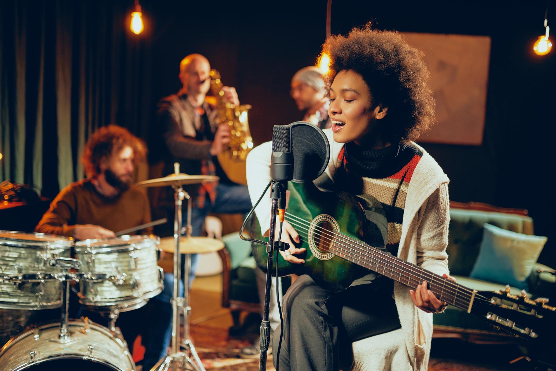 A woman sings into a microphone while playing guitar, with a drummer and saxophonist in the background on a dimly lit stage.