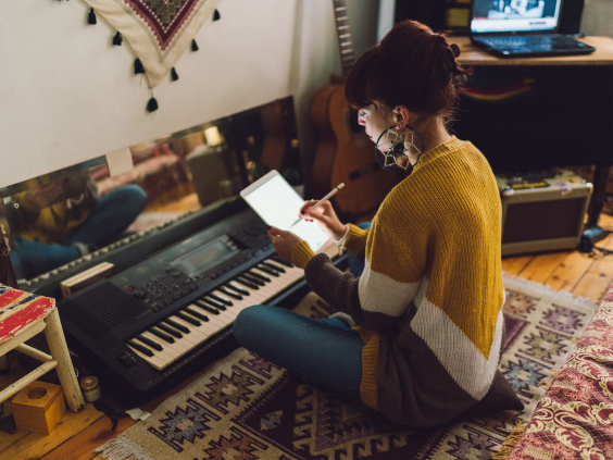A person in a cozy room sits on the floor writing on a tablet beside a keyboard, with a guitar and laptop nearby.