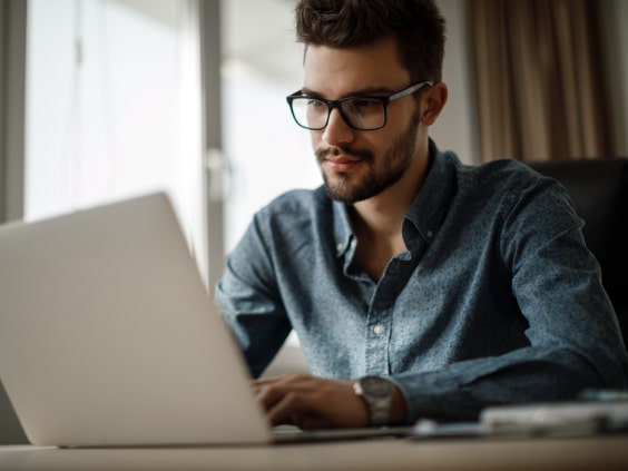 Mann mit Brille sitzt am Tisch und arbeitet am Laptop.