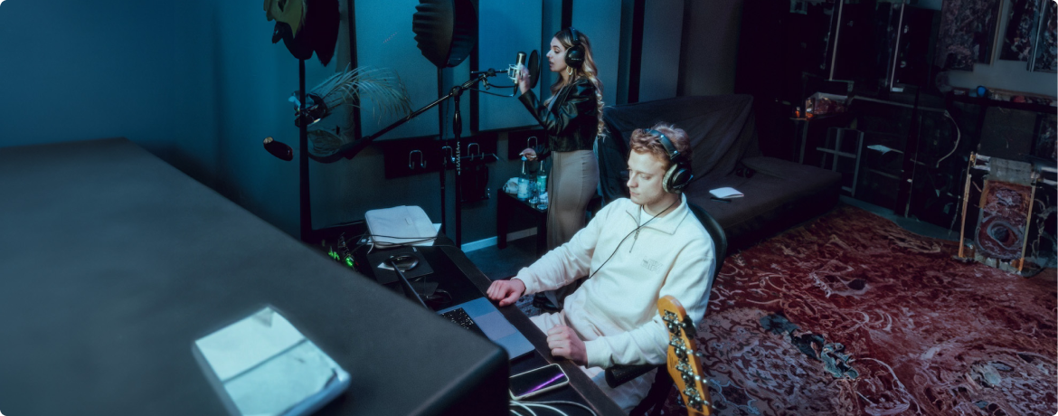 A woman sings into a microphone while a man works on a laptop in a recording studio with a red patterned carpet.
