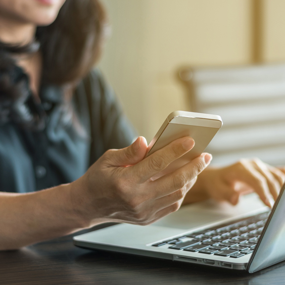 Frau sitzt am Tisch am Laptop mit einem Smartphone in der Hand.